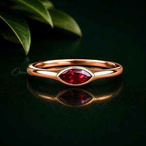 Könnte beinhalten: Ein Ring aus Roségold mit einem einzelnen, horizontal gefassten, marquise-geschliffenen roten Rubin in einer glatten Zargenfassung. Die Ringschiene ist poliert und massiv und zeigt ein schlichtes Design mit einem leuchtend roten Stein.