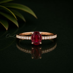 Puede incluir: Un anillo de oro rosa con un rubí rojo central de talla ovalada sujeto por cuatro garras. La banda está adornada con una hilera de pequeños diamantes blancos redondos engastados a ambos lados de la piedra central, creando un aspecto refinado.