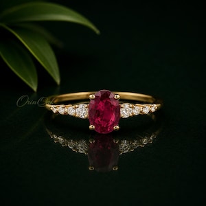 May include: A gold ring featuring a central oval-cut deep red ruby gemstone, flanked by three small round-cut white diamonds on each side of the band. The ring is set on a dark reflective surface with a green leaf visible in the upper left corner.