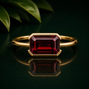 Könnte beinhalten: Ein Goldring mit einem horizontalen, smaragdgeschliffenen, tiefroten Edelstein in einer Zargenfassung. Die Ringschiene ist dünn und poliert und weist eine glatte metallische Oberfläche auf. Der Edelstein ist rechteckig mit facettierten Kanten.