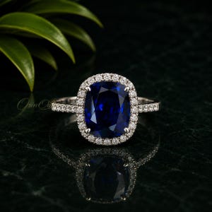 May include: A silver-toned ring featuring a large, cushion-cut deep blue sapphire center stone surrounded by a halo of small, clear round-cut diamonds. The band is also set with small clear diamonds along the sides, creating a brilliant sparkle.
