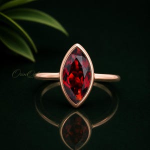 Puede incluir: Un anillo de oro rosa con una piedra preciosa granate de color rojo intenso en talla marquesa, montada en un engaste de bisel liso. La banda es fina y pulida, mostrando una piedra vibrante y facetada que refleja la luz con simetría.