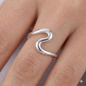 Op de afbeelding: Een gepolijste zilveren ring met een doorlopend, golfachtig ontwerp dat over de vinger buigt. De band heeft een glad, reflecterend metalen oppervlak met een vloeiende, golvende vorm die een open, asymmetrisch silhouet creëert.