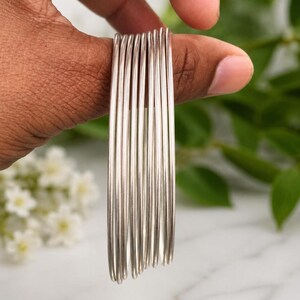 May include: A set of seven thin, polished silver-toned metal bangles stacked together. Each bangle features a smooth, rounded wire construction with a reflective metallic finish. The bracelets are arranged in a parallel vertical alignment.