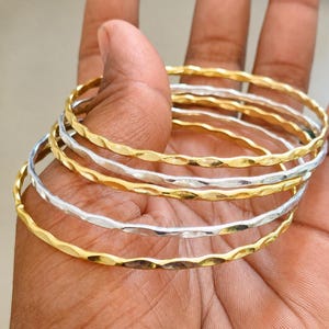 May include: A set of five thin, circular bangles featuring a repeating hammered texture. The collection includes alternating gold-toned and silver-toned metal bracelets, each with a consistent wavy, indented surface pattern throughout the entire band.