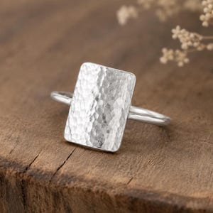 Könnte beinhalten: Ein Silberring mit einer großen, rechteckigen Platte, die eine gehämmerte, texturierte Oberfläche aufweist. Die Ringschiene ist schmal und glatt und mit der Rückseite der rechteckigen Platte verbunden. Das Metall glänzt hell auf der Oberfläche.