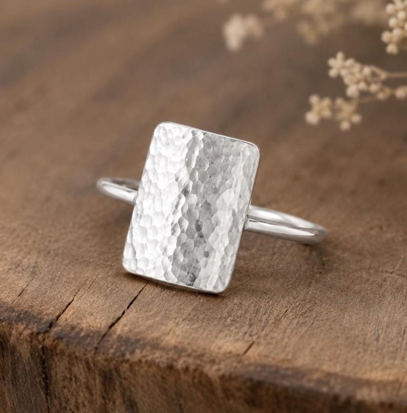 May include: A silver ring featuring a large, rectangular face with a hammered, textured finish. The band is thin and smooth, connecting to the back of the rectangular plate. The metal has a bright, reflective sheen across the entire surface.
