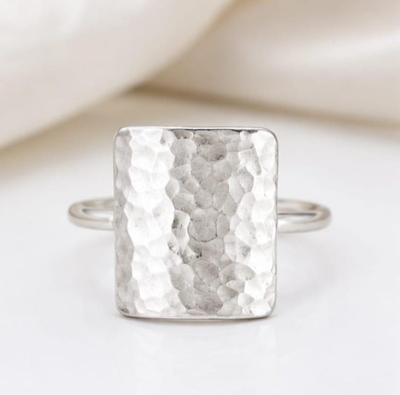 May include: A silver-toned ring featuring a large, rectangular face with a hammered, textured surface. The band is thin and smooth, connecting to the sides of the prominent, vertically oriented rectangular centerpiece with a reflective finish.