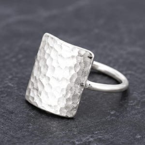 May include: A silver ring featuring a large, square-shaped face with a hammered, textured surface. The band is smooth and rounded, connecting to the back of the square plate. The metal has a bright, reflective finish throughout the entire piece.