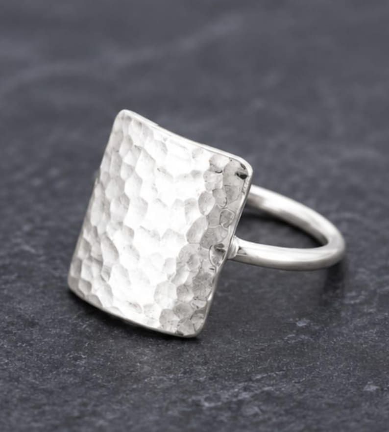 May include: A silver ring featuring a large, square-shaped face with a hammered, textured surface. The band is smooth and rounded, connecting to the back of the square plate. The metal has a bright, reflective finish throughout the entire piece.