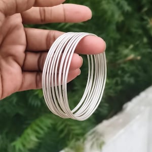 May include: A set of multiple thin, smooth, silver-coloured metal bangles stacked together. Each bangle features a simple, rounded wire design with a polished finish, creating a uniform and cohesive look when worn as a single layered accessory.