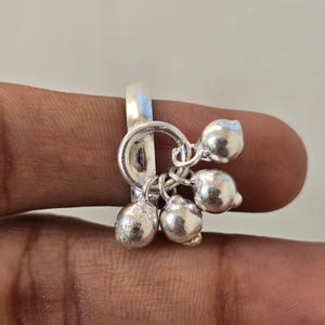 May include: A silver-toned ring featuring a smooth, solid band and a circular charm attachment. Three small, spherical, bell-like charms hang from the central ring, each with a polished, reflective metallic finish and a rounded, bulbous shape.