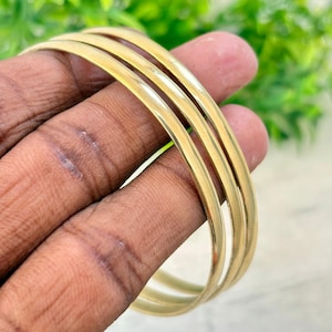 May include: A set of three smooth, polished gold-tone metal bangles. Each bracelet features a simple, rounded, continuous circular design with a reflective surface. The bangles are stacked together, showcasing a uniform metallic yellow finish.