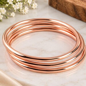 Può includere: Un set di tre bracciali rigidi in metallo color oro rosa lucido. Ogni bracciale presenta un design a fascia circolare continua, liscia e arrotondata, con una finitura riflettente e brillante. I tre bracciali singoli sono impilati insieme.