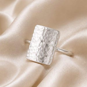 May include: A silver-coloured ring featuring a large, rectangular face with a hammered, textured surface. The band is thin and smooth, connecting to the sides of the rectangular centrepiece. The metal has a reflective, polished finish throughout.