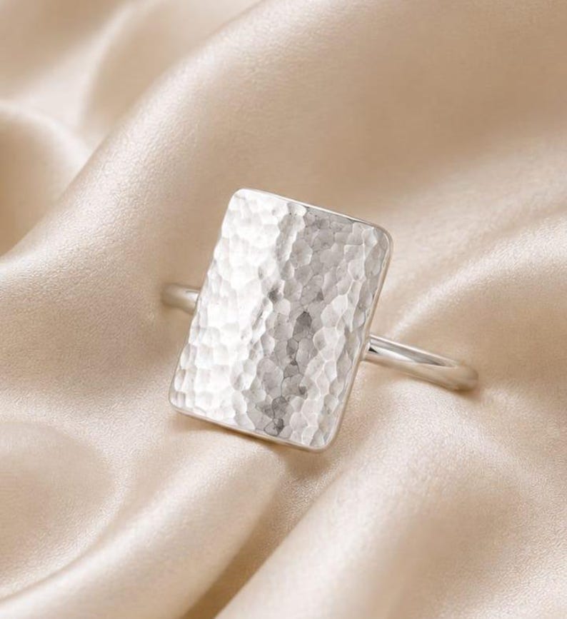 May include: A silver-coloured ring featuring a large, rectangular face with a hammered, textured surface. The band is thin and smooth, connecting to the sides of the rectangular centrepiece. The metal has a reflective, polished finish throughout.