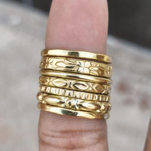 Könnte beinhalten: Ein Stapel aus sieben goldfarbenen Metallringen an einem Finger. Die Ringe weisen verschiedene Texturen und Muster auf, darunter glatte Bänder, blumenartige Gravuren, sich wiederholende ovale Formen, vertikale Rillen und längliche Perlen.
