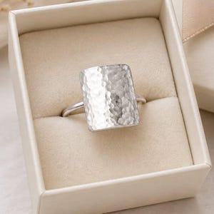 May include: A silver-toned ring featuring a large, rectangular face with a hammered, textured surface. The ring is presented inside a square, cream-coloured gift box with a soft, padded interior lining and a matching ribbon detail on the corner.