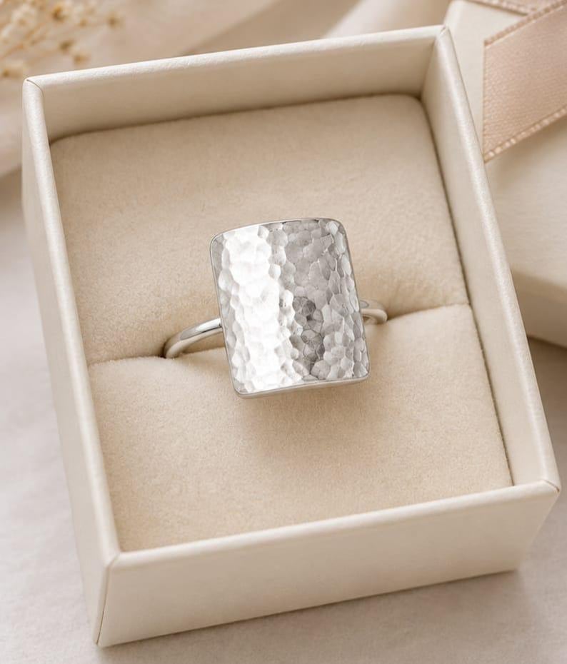 May include: A silver-toned ring featuring a large, rectangular face with a hammered, textured surface. The ring is presented inside a square, cream-coloured gift box with a soft, padded interior lining and a matching ribbon detail on the corner.