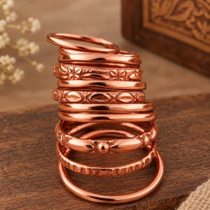 Op de afbeelding: Een stapel van tien koperkleurige metalen ringen. De collectie bevat verschillende gladde banden naast ringen met diverse reliëfpatronen, zoals bloemmotieven, geometrische vormen en kraalachtige texturen, allemaal gerangschikt in een verticale kolom.