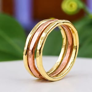 May include: A tri-color ring featuring three interlocking bands with a hammered texture. The design consists of one central band in a copper tone flanked by two outer bands in a bright yellow gold tone, creating a layered and reflective appearance.