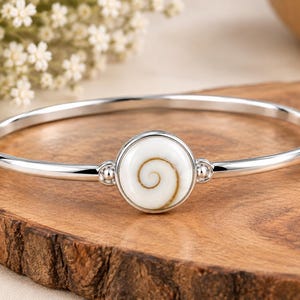 Puede incluir: Una pulsera rígida de plata con un engaste circular central que contiene una concha de ojo de shiva blanca con un patrón de espiral marrón natural. La banda de metal pulido es lisa y se conecta al punto focal con pequeñas cuentas de plata.