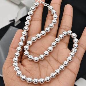 May include: A polished silver bead necklace featuring a continuous strand of uniform, reflective spherical beads. The necklace is secured with a small, circular silver clasp mechanism that connects the two ends of the beaded chain together.