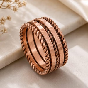 May include: A wide copper ring featuring a central band with a hammered texture, flanked on both sides by two twisted rope-style bands. The metallic surface has a warm, reddish-brown tone with a polished finish, creating a layered appearance.