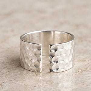 Könnte beinhalten: Ein breiter Silberring mit offenem Banddesign und einer texturierten, gehämmerten Oberfläche. Das Metall weist zahlreiche kleine, kreisförmige Vertiefungen auf, die das Licht reflektieren und dem Ring ein unebenes, glänzendes Aussehen verleihen.