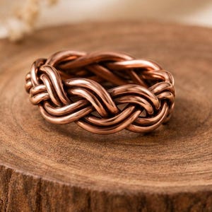 May include: A polished copper ring featuring a continuous woven or braided design. The metal has a warm, reddish-brown metallic luster with smooth, rounded strands that interlock to form a circular band with a consistent, repeating pattern.