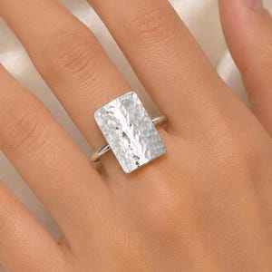 May include: A silver-toned ring featuring a large, rectangular face with a hammered, textured surface. The metal band is thin and connects to the back of the rectangular plate, creating a bold, reflective piece of jewellery worn on a finger.