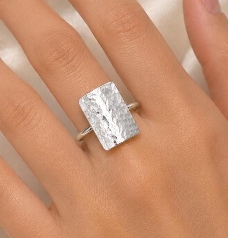 May include: A silver-toned ring featuring a large, rectangular face with a hammered, textured surface. The metal band is thin and connects to the back of the rectangular plate, creating a bold, reflective piece of jewellery worn on a finger.
