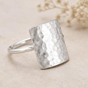 May include: A silver ring featuring a large, rectangular face with a hammered, textured surface. The band is smooth and polished, connecting to the sides of the rectangular plate. The metal has a bright, reflective finish throughout the piece.