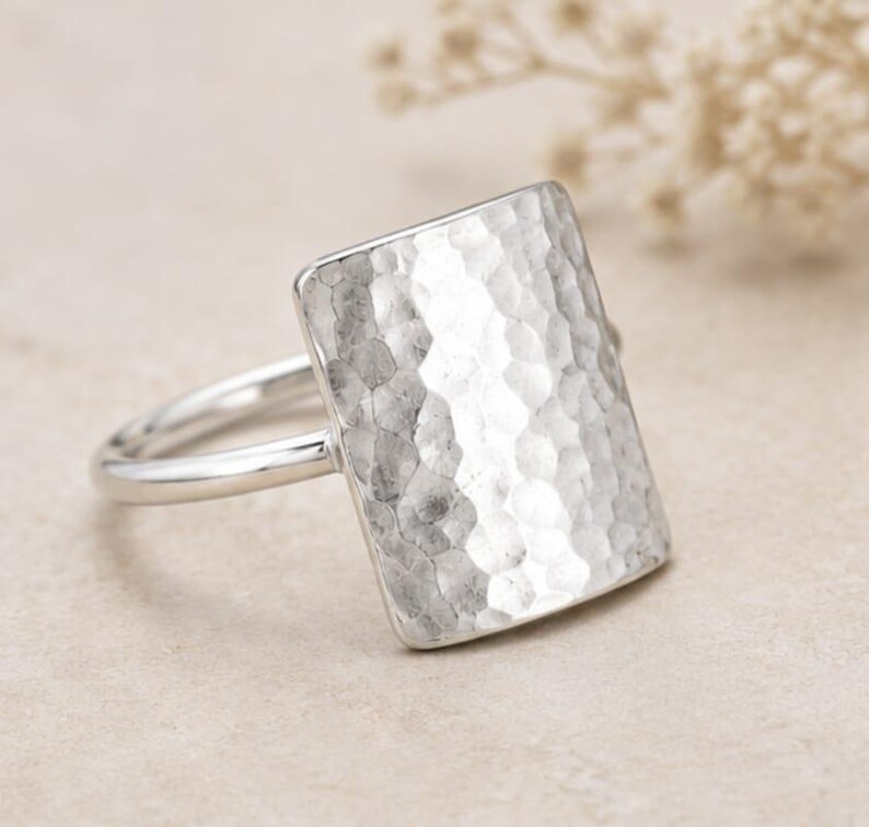 May include: A silver ring featuring a large, rectangular face with a hammered, textured surface. The band is smooth and polished, connecting to the sides of the rectangular plate. The metal has a bright, reflective finish throughout the piece.