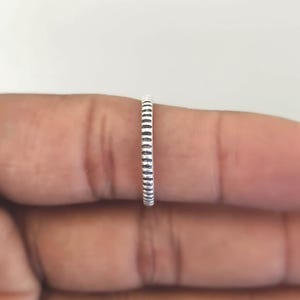 May include: A thin silver ring featuring a repeating vertical line pattern along the entire outer band. The metal has a slightly oxidized finish within the grooves, creating a contrast between the raised silver ridges and the dark recessed lines.