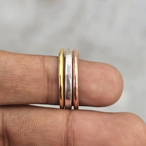 Pode incluir: Um conjunto de três anéis de metal finos e lisos empilhados. Os anéis apresentam acabamentos metálicos distintos, incluindo uma banda em tom de ouro amarelo, uma banda em tom prateado e uma banda em tom de cobre ou ouro rosa.