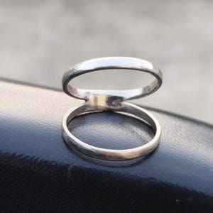 Puede incluir: Un anillo de color plateado con un diseño de doble banda. El anillo consta de dos bandas circulares finas y lisas conectadas en un solo punto, creando una apariencia abierta y superpuesta con un acabado metálico pulido y brillante.