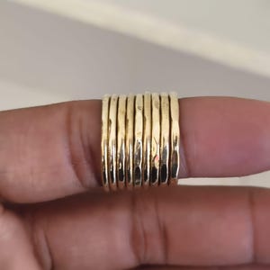 May include: A set of eight thin, gold-colored metal rings with a hammered, textured surface. The rings are stacked side-by-side, featuring a consistent metallic finish and a rounded, slender band design that creates a uniform appearance across the set.