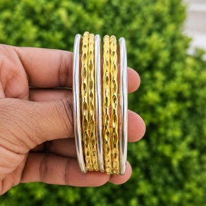Op de afbeelding: Een set van zes gestapelde armbanden met afwisselende ontwerpen. Twee gladde zilverkleurige armbanden flankeren twee getextureerde goudkleurige armbanden in het midden, met twee extra gladde zilverkleurige armbanden aan de buitenranden.