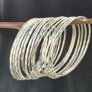 Puede incluir: Un conjunto de diez brazaletes finos de color plateado con un acabado martillado y texturizado. Las pulseras circulares están dispuestas en fila, reflejando la luz en sus superficies metálicas, creando un patrón repetitivo de líneas.