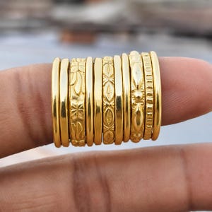 Pode incluir: Um conjunto de dez anéis de metal em tom dourado empilhados. A coleção inclui várias bandas lisas e simples, além de anéis com padrões em relevo intrincados, incluindo motivos florais, formas geométricas e linhas verticais texturizadas.