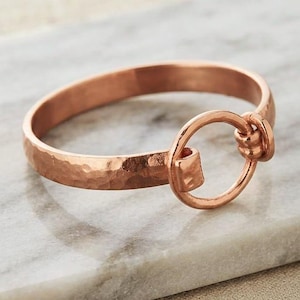 Peut inclure: Un bracelet en métal de couleur cuivre présentant une texture martelée sur la bande. Le bracelet est conçu avec un élément circulaire en forme d'anneau fixé à une extrémité, servant de mécanisme de fermeture relié à l'autre côté de la bande.