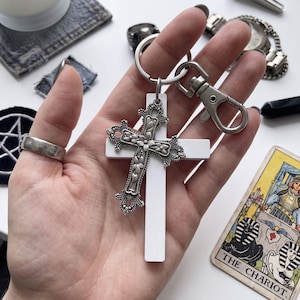 Può includere: Un portachiavi con una croce bianca semplice sovrapposta a una croce in metallo color argento più piccola e decorata con dettagli floreali. La croce è fissata a un anello diviso color argento e a un moschettone per un fissaggio sicuro.