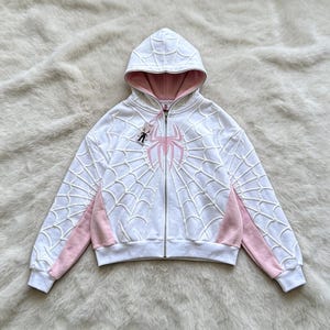 Op de afbeelding: Een wit vest met capuchon en rits, voorzien van een verhoogd, gestructureerd spinnenwebpatroon op de voor- en achterkant en de mouwen. Het kledingstuk heeft een lichtroze spinnenembleem op de borst, een roze voering en roze zijpanelen.