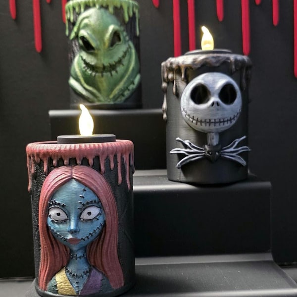 Könnte beinhalten: Drei zylindrische LED-Kerzen mit skulptierten Figuren aus Nightmare Before Christmas. Eine Kerze zeigt den grünen Oogie Boogie, eine den weißen Jack Skellington und eine die blauhäutige Sally mit langem rotem Haar.