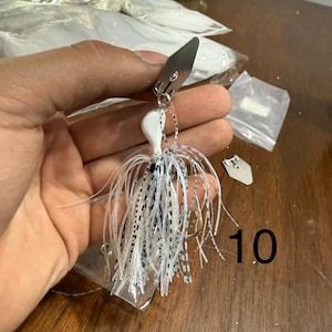 May include: A fishing lure featuring a white jig head and a skirt made of thin, white and black speckled silicone strands. A silver-colored metal blade is attached to the top of the jig head by a small metal ring. The number 10 is visible.