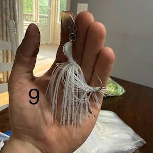 May include: A fishing lure featuring a white head, a silver metal blade, and a skirt made of thin, translucent white silicone strands with small black glitter flecks. The number 9 is printed in black on the left side of the image.