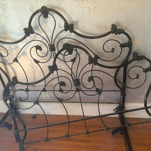 May include: A black metal bed frame featuring an ornate headboard and footboard with intricate scrollwork, curved iron bars, and decorative floral-shaped accents at the joints. The structure is composed of thin, dark metal rods arranged in a symmetrical pattern.