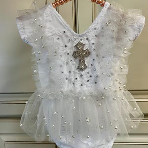 May include: A white lace romper featuring ruffled sleeves and a tulle skirt overlay. The garment is embellished with scattered white pearls and silver sequins, and it features a prominent central cross applique adorned with clear crystals.