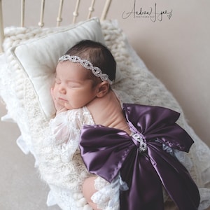 May include: A newborn infant wearing a white lace wrap with a large, deep purple satin bow on the back and a silver-toned rhinestone headband. The infant is resting on a cream-colored chunky knit blanket inside a small, vintage-style metal bed frame.